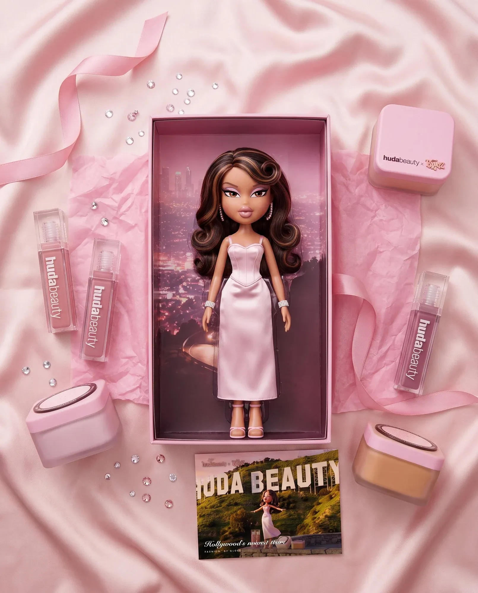 The PR kit laid out on pink satin: the doll, both glosses, both powders and the card
