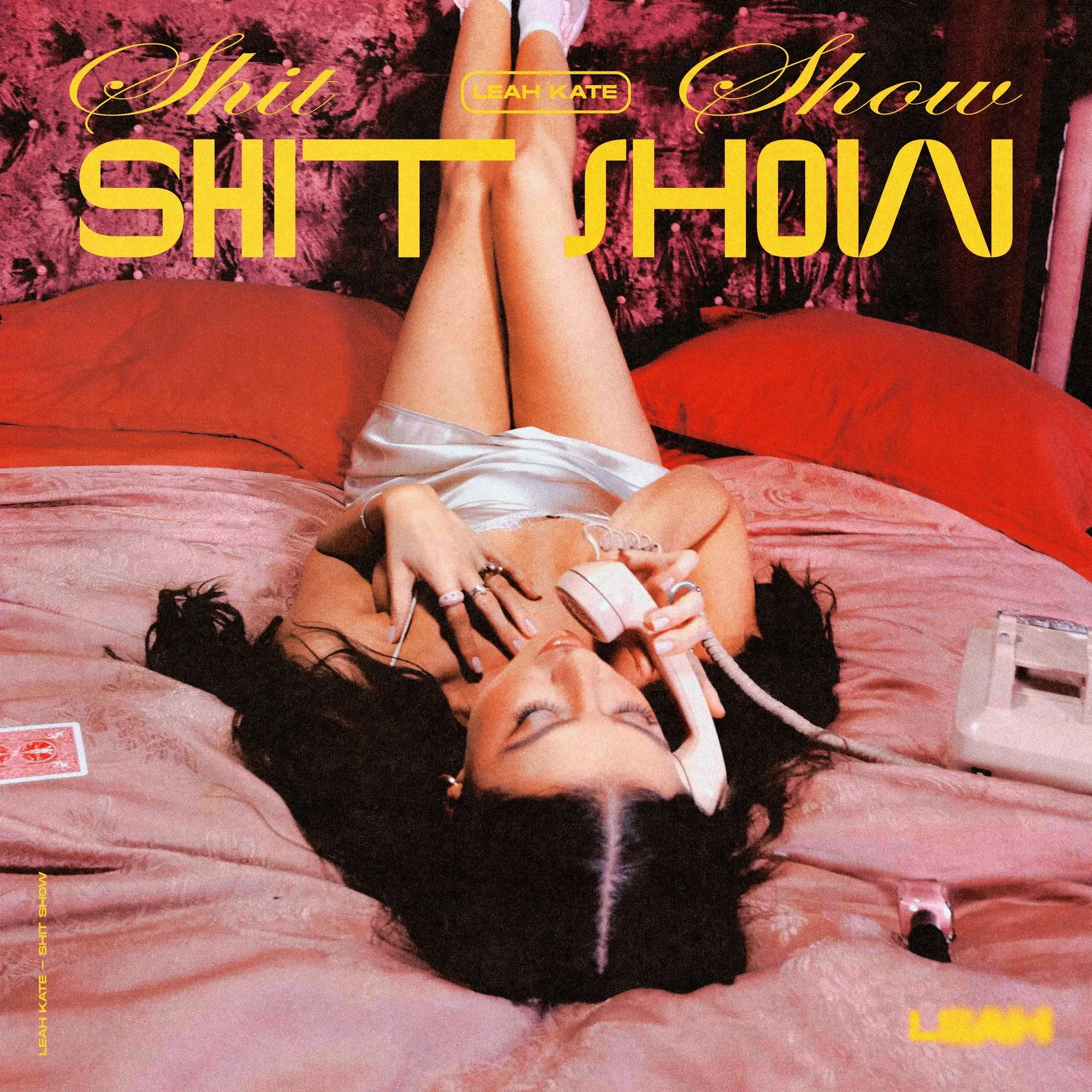 Shit Show — a hot pink and red bedroom, Leah Kate upside down on the bed with a corded phone, playing cards scattered, the title in acid yellow