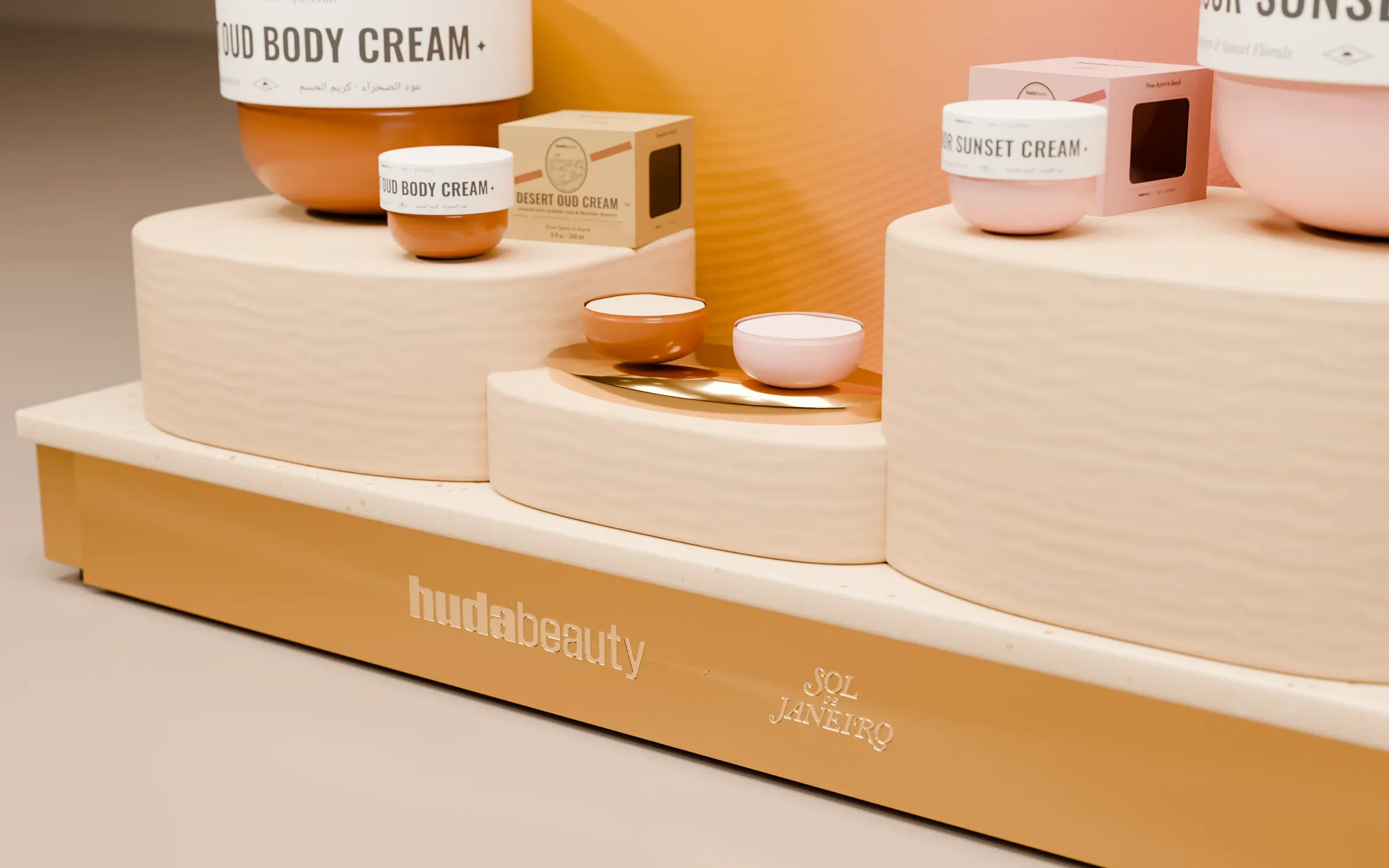 Close-up: dune-textured plinths, tester tray and the engraved huda beauty × Sol de Janeiro base